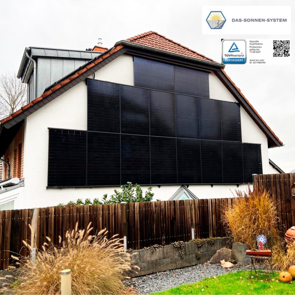 Ein Haus mit schwarzen Solarpanelen an der Fassade, umgeben von Herbstpflanzen und einem Zaun.